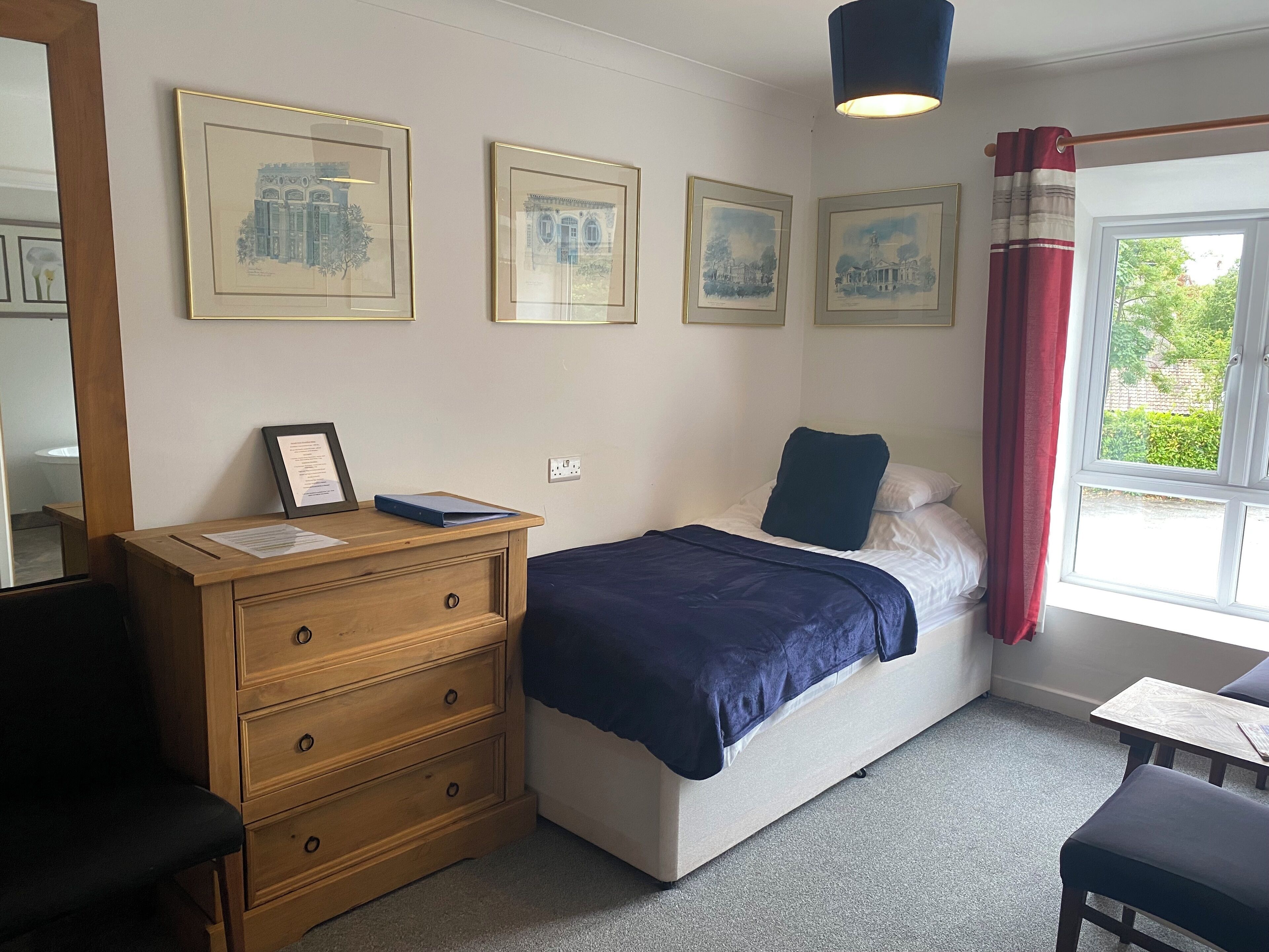 A bedroom with a bed, a wooden dresser, a desk, and a window with curtains.