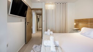 A hotel room with a bed, a TV mounted on the wall, a bedside table with a lamp, and a bathroom visible through an open door. - Directly before the sea.Direkt vor dem Meer (Vouliagmeni)
