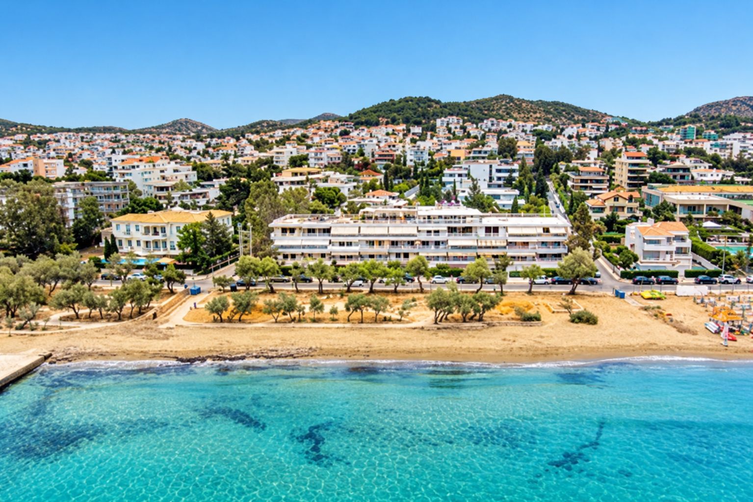 A coastal town with a beachfront hotel featuring multiple floors, a sandy beach, and clear blue waters.