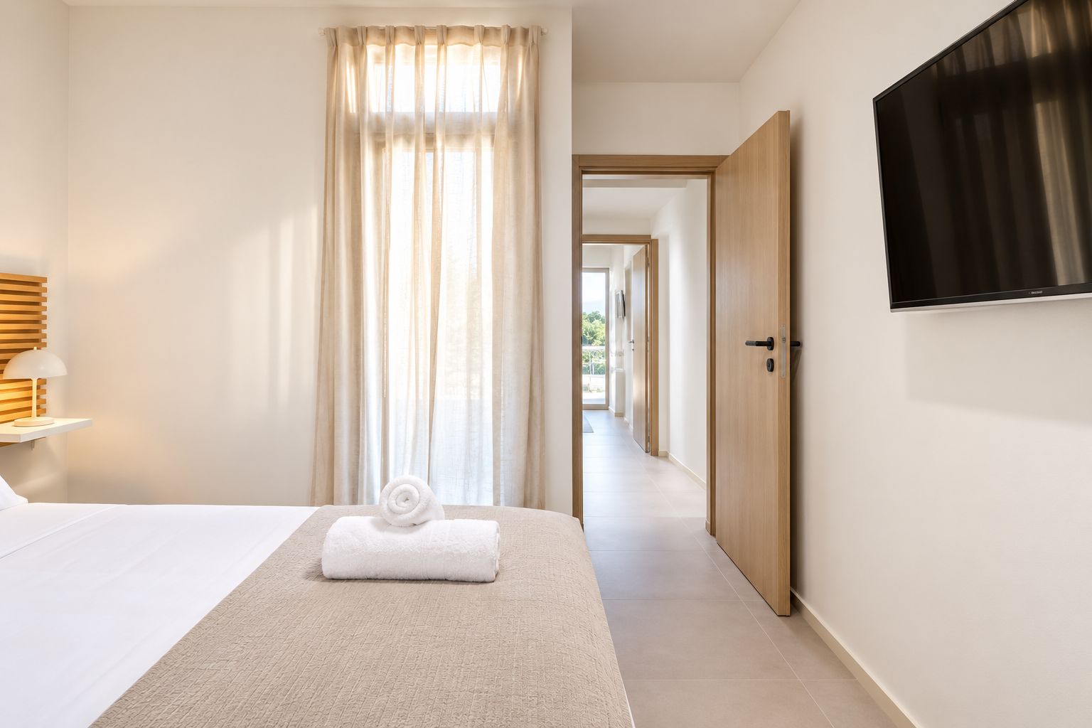 A hotel room with a bed, a TV, a window with curtains, a door, and a hallway leading to another room.