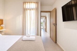 A hotel room with a bed, a TV, a window with curtains, a door, and a hallway leading to another room. - Directly before the sea.Direkt vor dem Meer (Vouliagmeni)