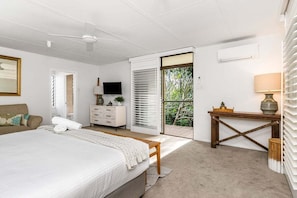 A bedroom with a bed, a dresser, a TV, a window with blinds, and a view of greenery outside.