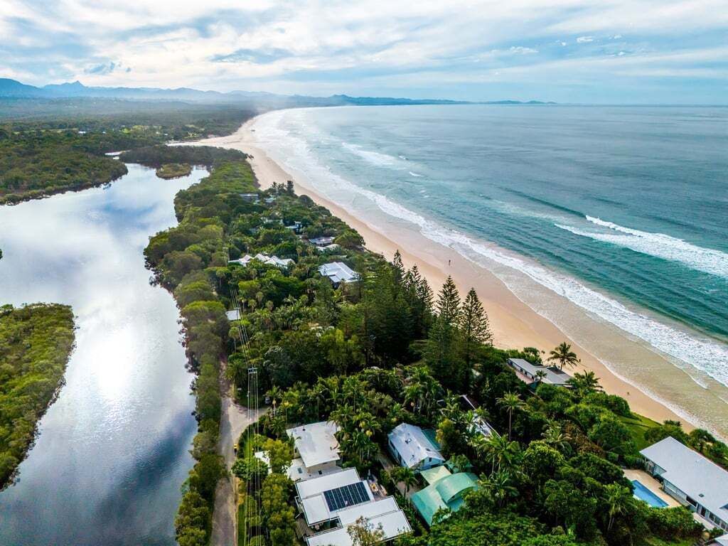 Vue aérienne d’une zone côtière comprenant une plage, une rivière et un ensemble de bâtiments entourés de verdure.