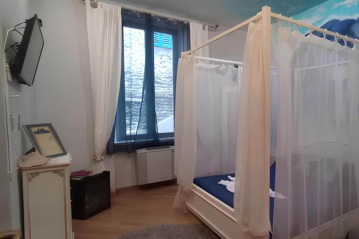 Quarto com cama de dossel, janela com cortinas, escrivaninha e televisão fixada na parede.