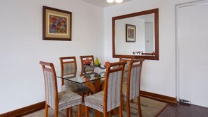 A dining area with a glass-top table, four striped chairs, a wooden side table with fruit, a framed picture, and a mirror on the wall. - Aparts4Days 2 Bedroom Apart San Isidro (Lima)