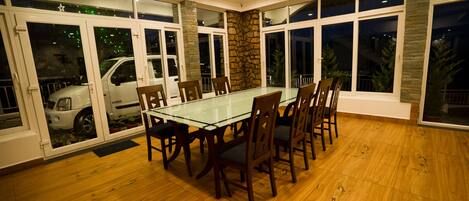 Een eetruimte met een glazen tafel, houten stoelen, grote ramen met uitzicht en een accentmuur van steen.