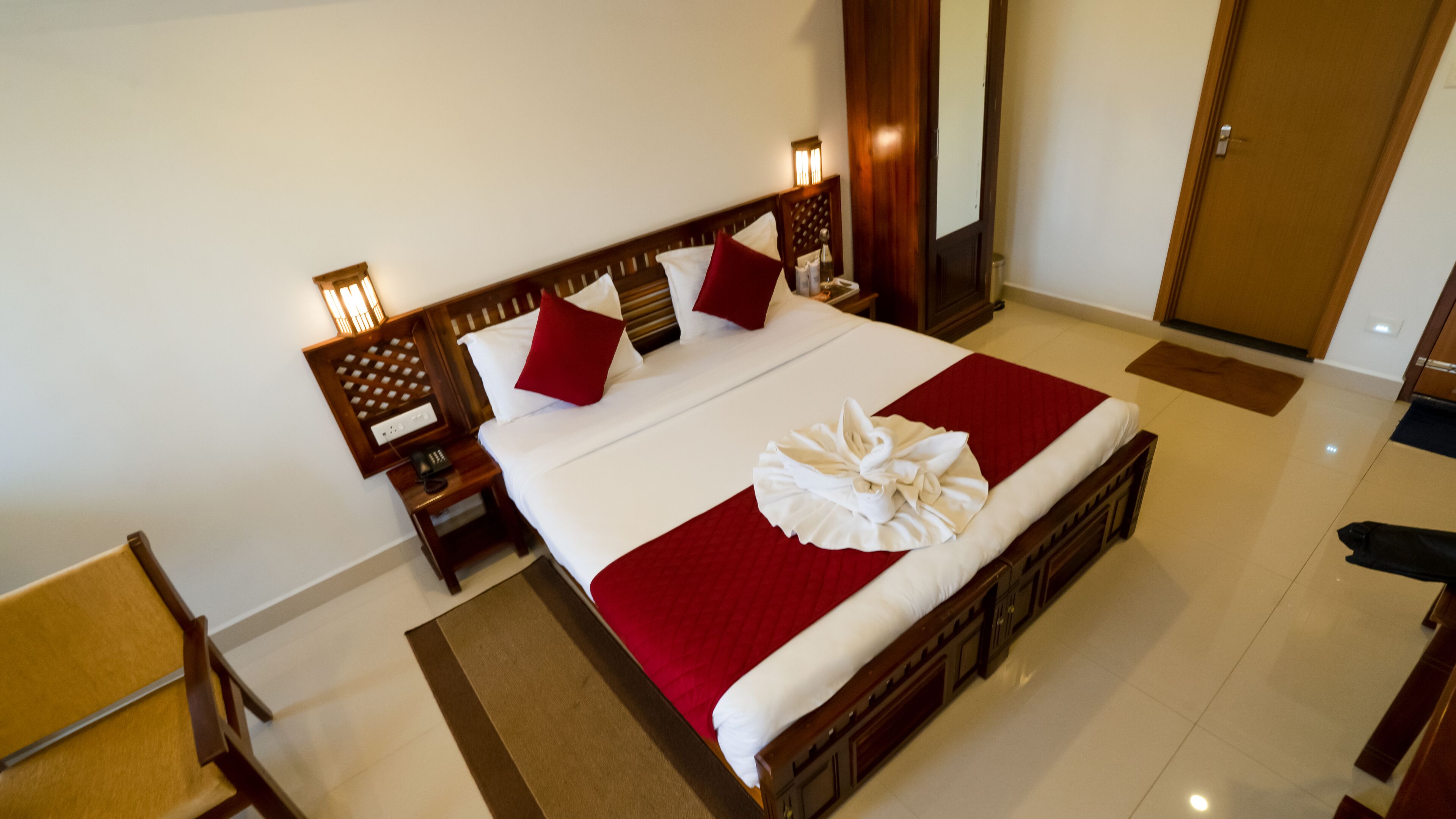 Habitación de hotel con una cama grande, cabecera de madera y un corredor rojo decorativo. Se ve un mesita de noche de madera con una lámpara y un teléfono. El piso es de baldosas y a la derecha hay una puerta.