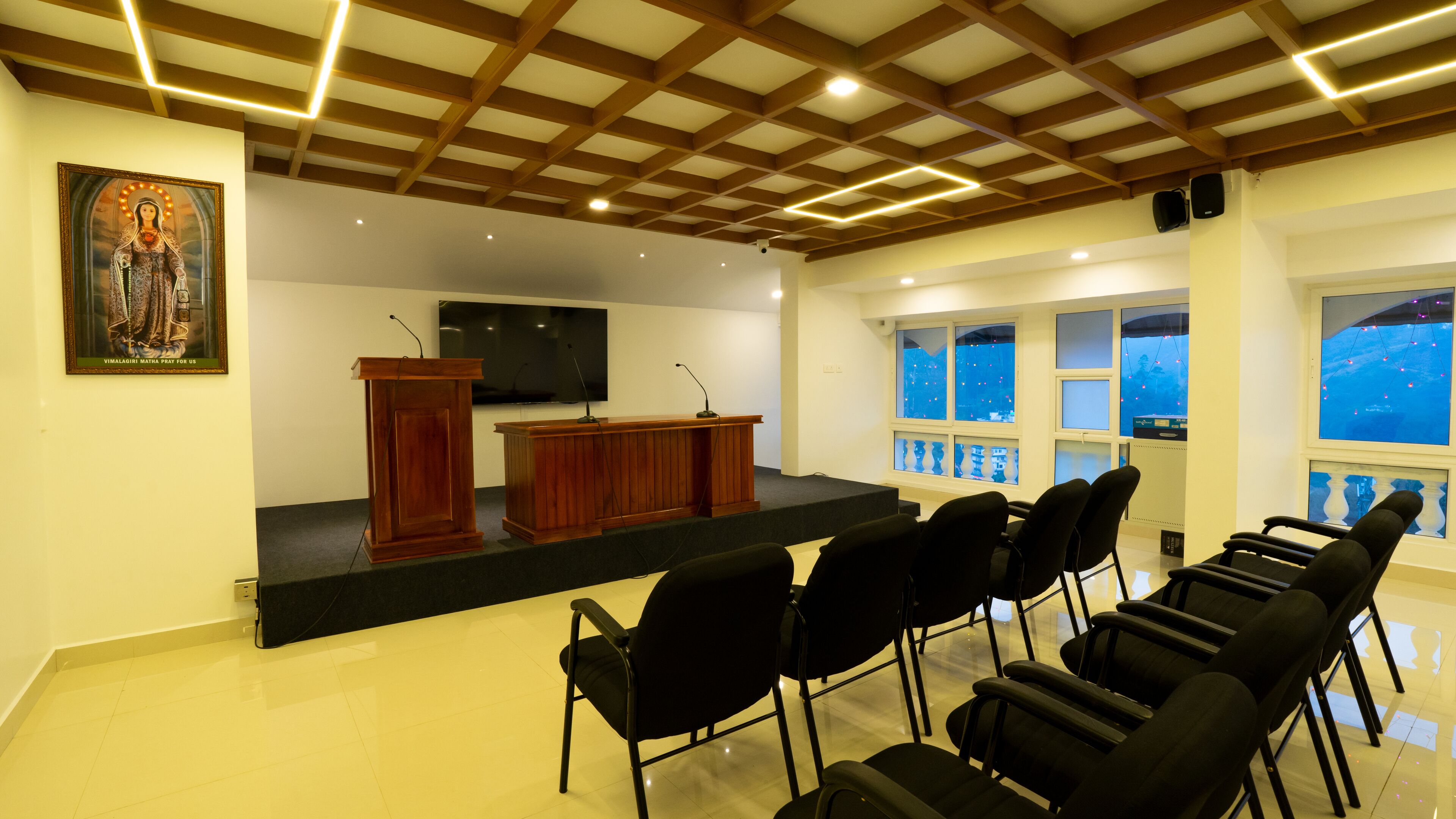 Ein Konferenzraum mit einem hölzernen Rednerpult, Stühlen, einer großen Leinwand und einem gerahmten religiösen Gemälde.