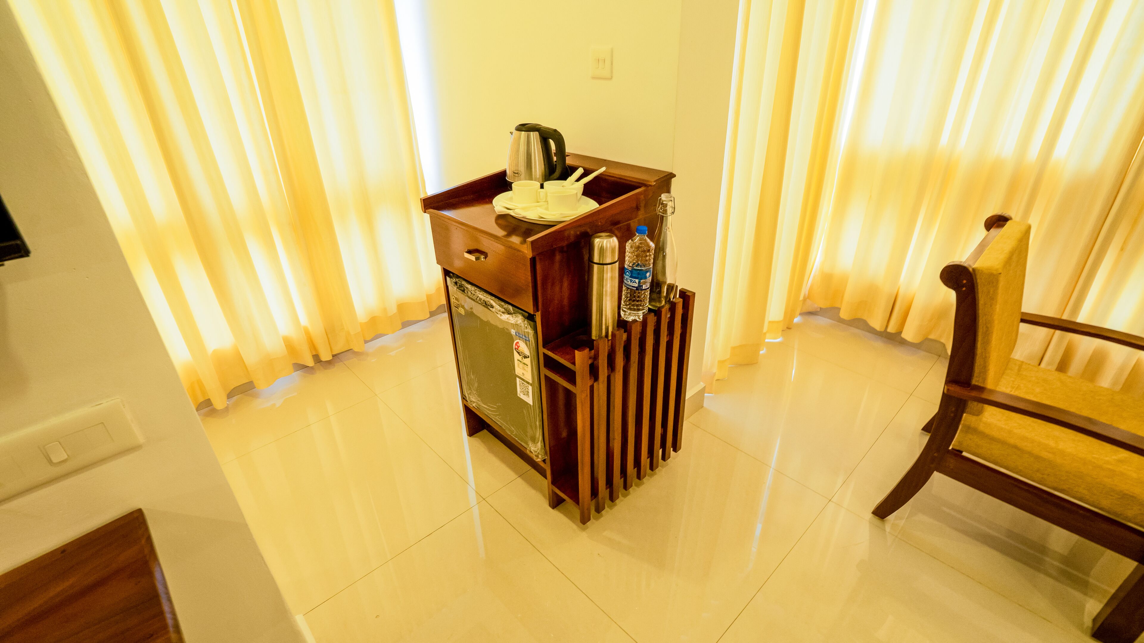 Habitación pequeña con un aparador de madera, un refrigerador, una silla y un ventanal con cortinas.
