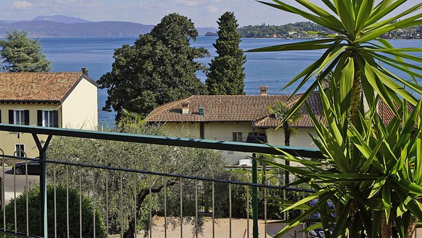 A balcony with a view of a lake, a potted plant, and a building with a tiled roof. - Blu View Apartment in Salo (Salò)
