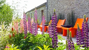 Un jardín con flores moradas y sillas anaranjadas frente a un edificio de piedra.