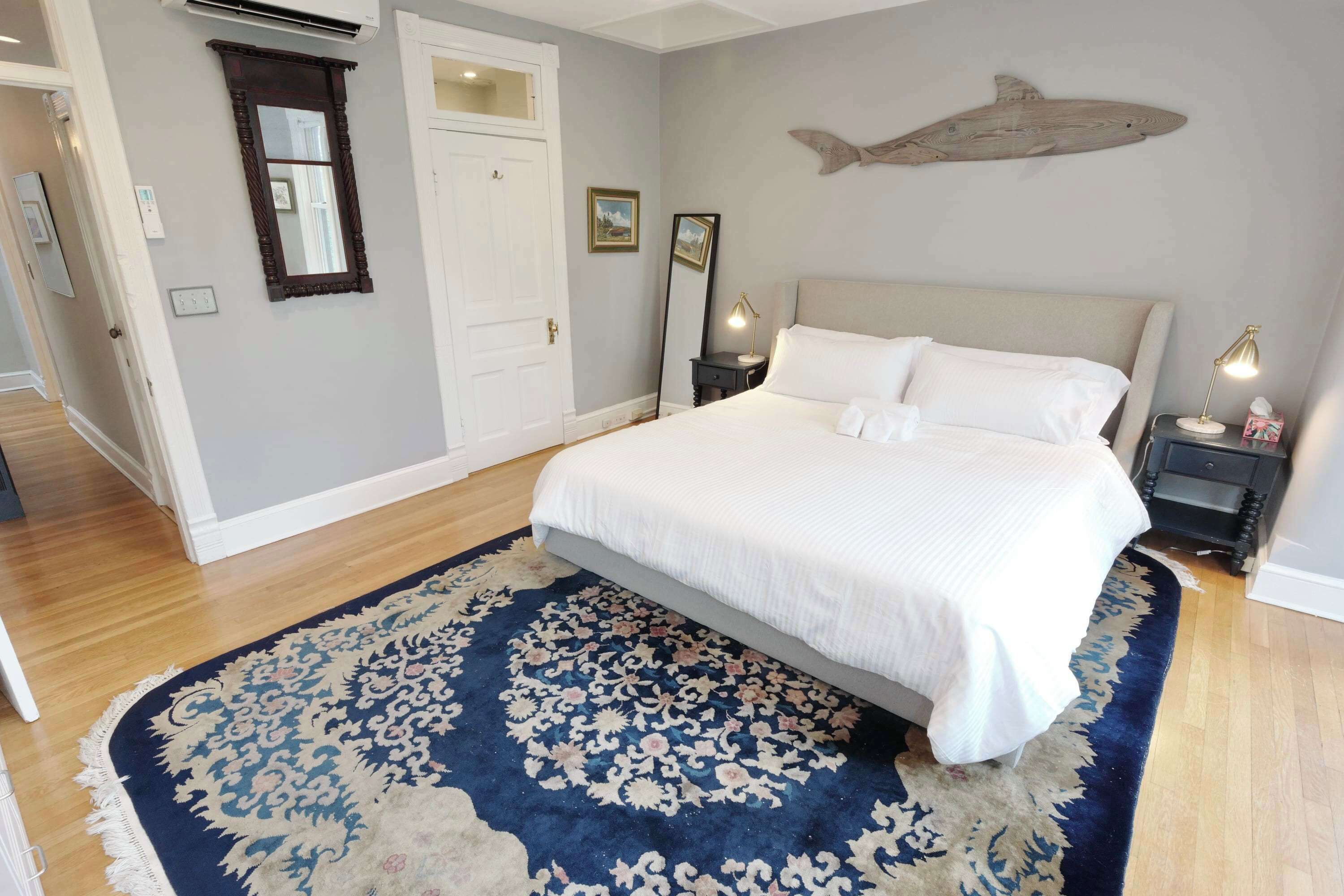 A bedroom with a bed, a large rug, a wall-mounted fish decoration, a door, and a nightstand with a lamp.