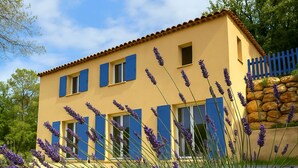 Ein zweistöckiges Gebäude mit gelber Fassade und blauen Fensterläden, umgeben von Lavendelpflanzen.