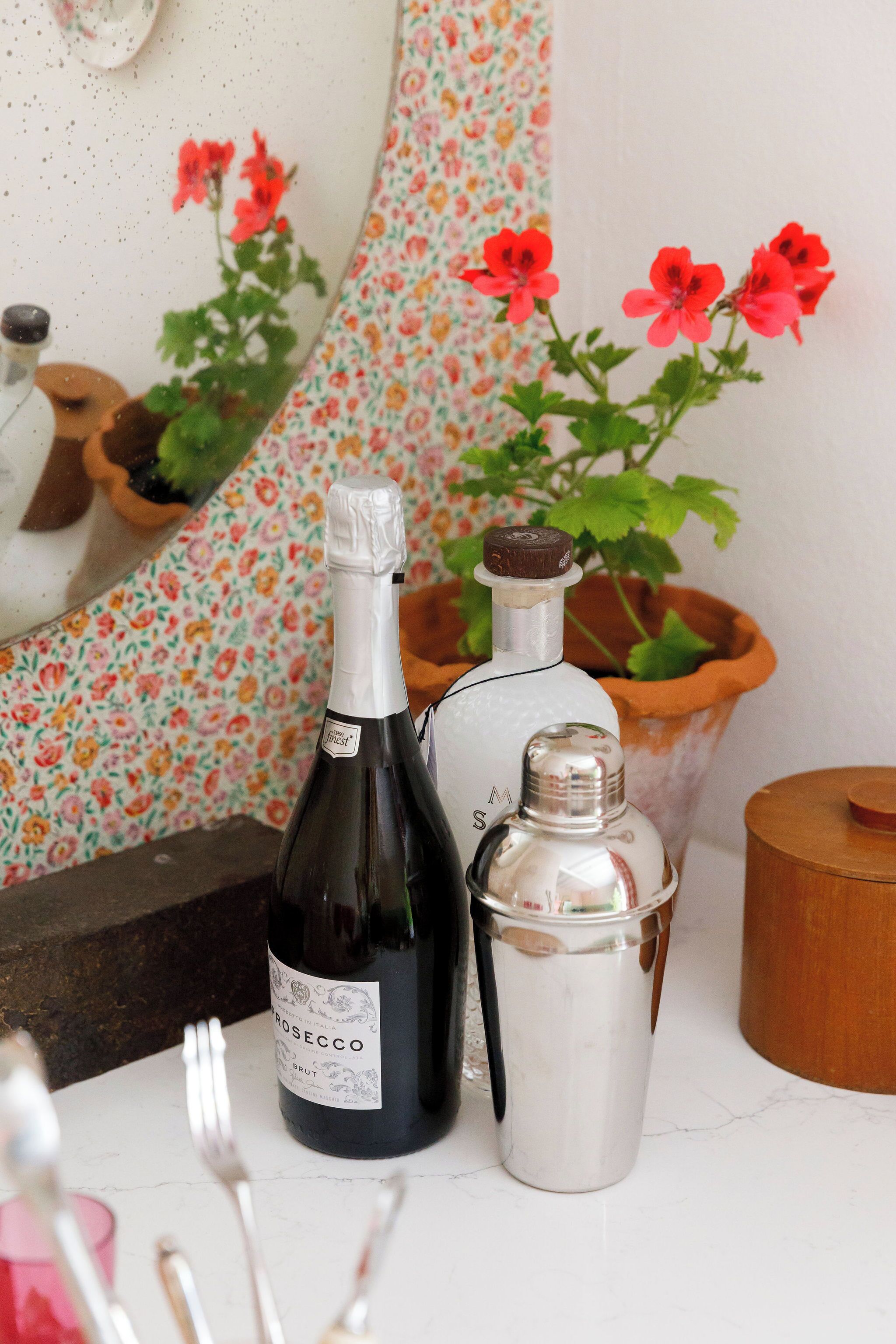 Une bouteille de vin rosé, un récipient en argent et une plante en pot avec des fleurs rouges, le tout posé sur une surface blanche.