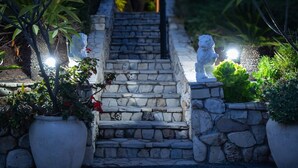Un escalier en pierre, orné de statues au sommet, entouré de plantes en pot et bordé d’un sentier.