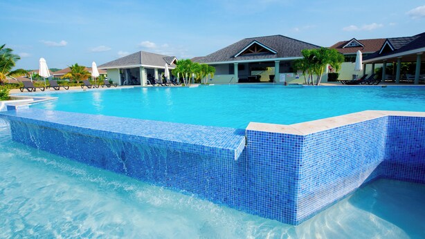 A clear blue swimming pool with lounge chairs and umbrellas, surrounded by buildings with a light-colored exterior and dark roofs.