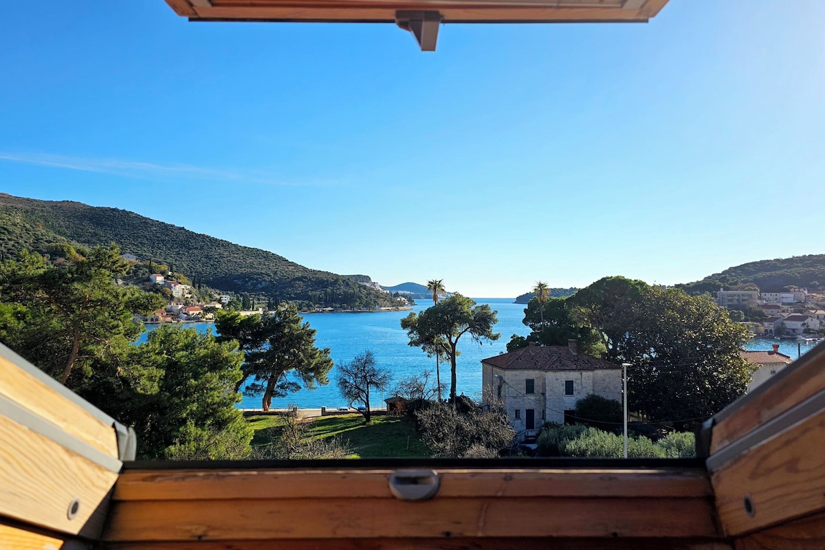 A view from a wooden balcony overlooking a coastal town with a clear blue sea, hills, and scattered buildings.