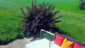 Ein Bett mit weißer Bettdecke und roten Kissen, ein kleiner Tisch mit Blumenvase sowie Blick auf ein grünes Feld und Bäume.