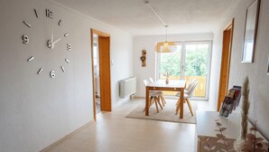 Ein moderner Essbereich mit Holztisch und -stühlen, einer großen Wanduhr und Blick ins Freie durch eine Glastür.