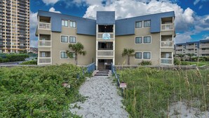 Un immeuble à plusieurs étages, doté d’une entrée centrale et de balcons, entouré de verdure et bordé d’un chemin de sable.