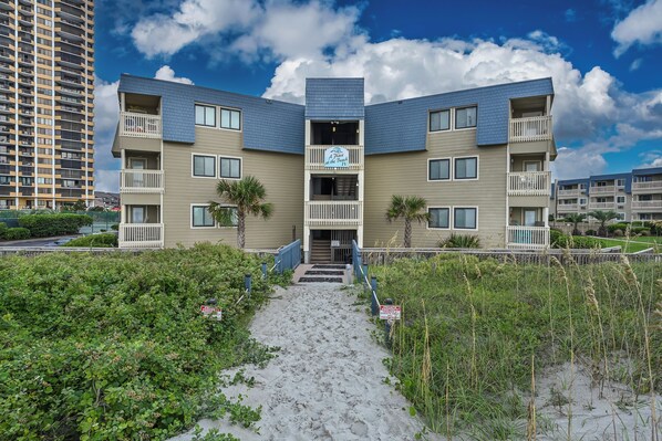 A multi-story building with a central entrance and balconies, surrounded by greenery and a sandy path. - Mermaid Cove (Horry County)