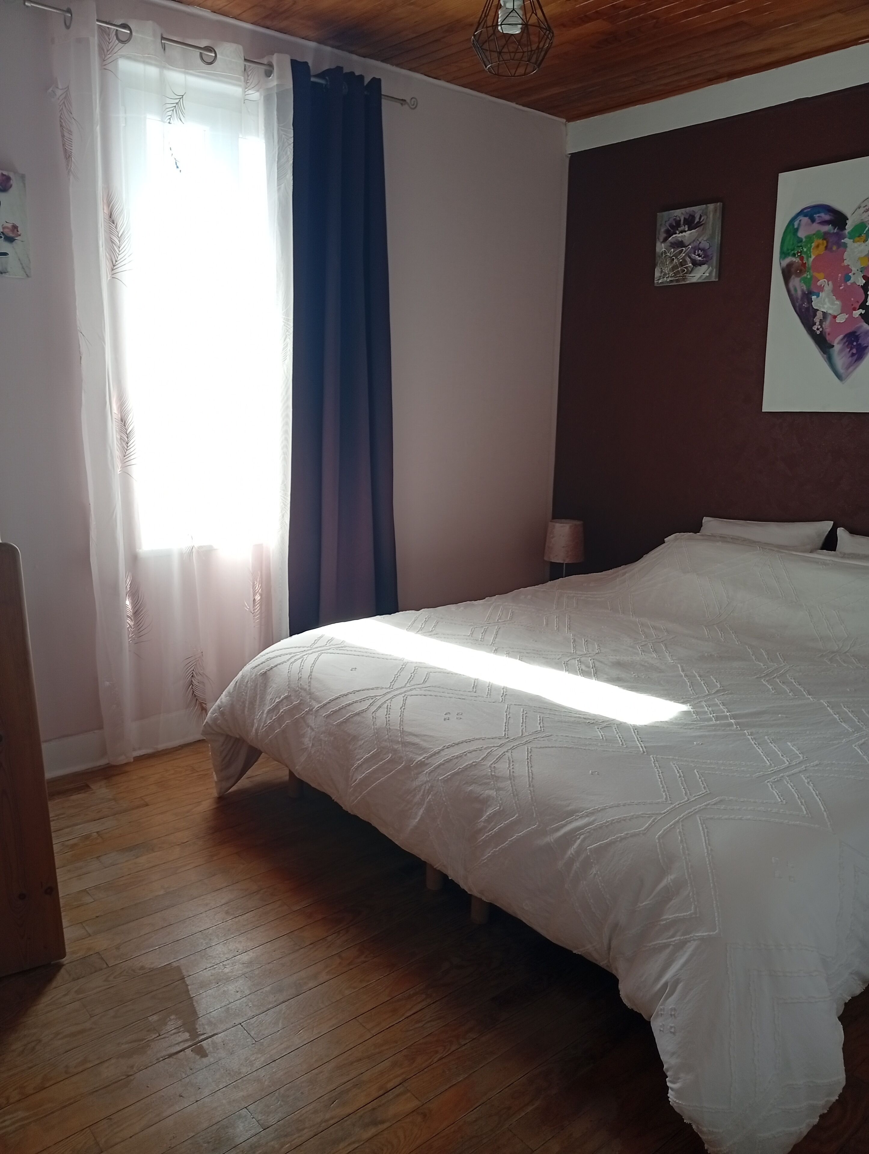 A bedroom with a bed, wooden floor, curtains, and a wall with artwork.
