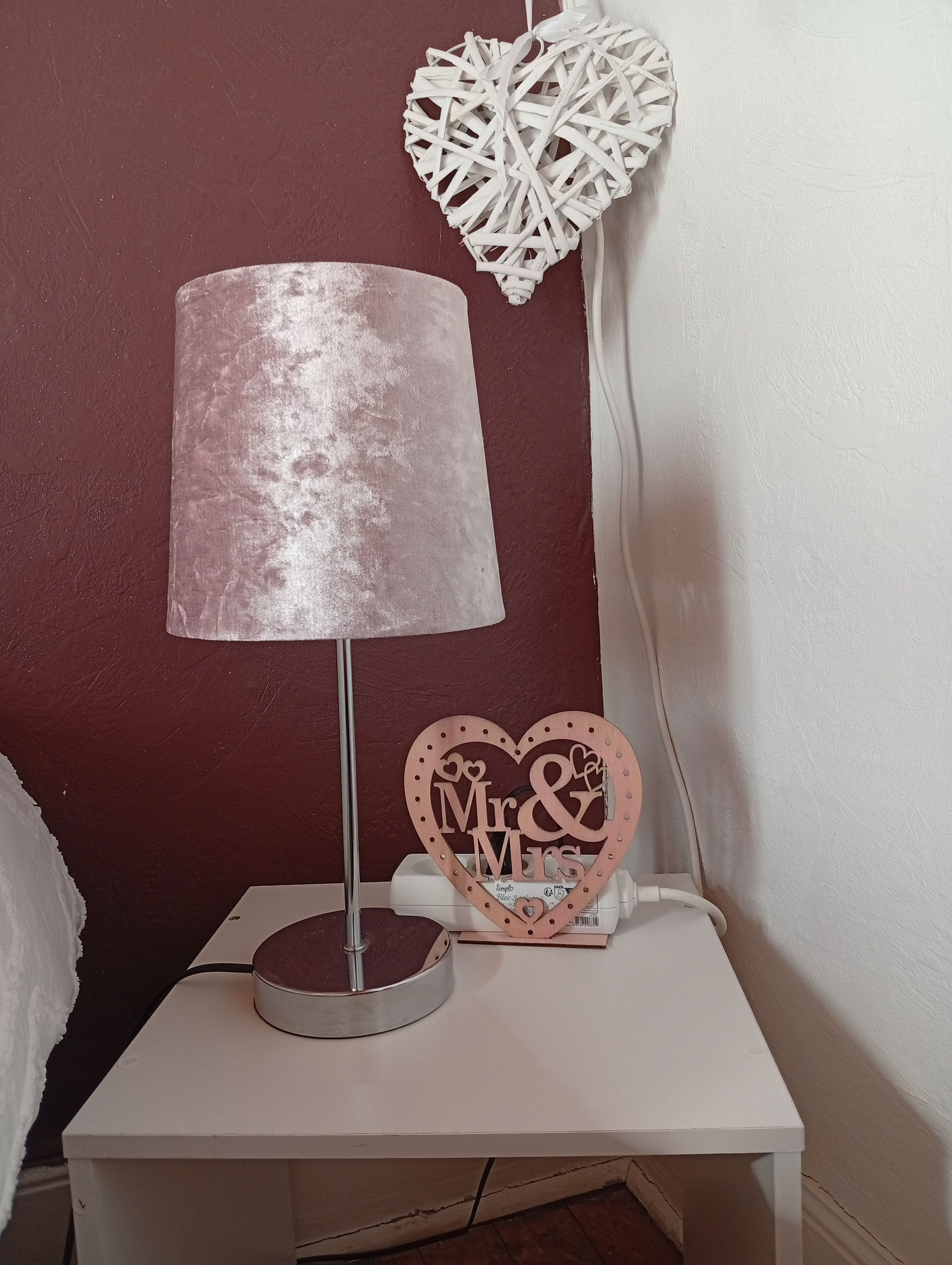 A bedside table with a lamp, a decorative heart-shaped sign, and a wall-mounted heart decoration.
