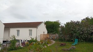 Ein weißes Haus mit einem Ziegeldach, ein Garten mit verschiedenen Pflanzen, ein Holzzaun und ein grüner Stuhl.