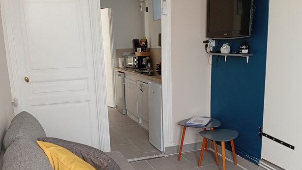 Ein Wohnzimmer mit grauem Sofa, einem gelben Kissen, einem kleinen runden Tisch mit Stuhl, einer Küche im Hintergrund und einem an einer blauen Wand montierten Fernseher.