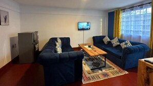 A compact living space with two blue sofas, a small wooden table, a television mounted on the wall, and a refrigerator.
