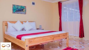 A bedroom with a wooden bed, white bedding, a red throw, a nightstand, a painting on the wall, and a window with red curtains.
