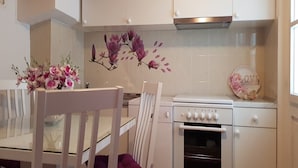 Una cocina con armarios blancos, una estufa y un horno empotrado. Hay una silla blanca y una mesa con un florero.