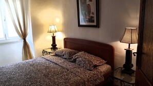Una cama bien hecha con ropa de cama estampada, una mesita de noche con una lámpara, un cuadro en la pared y una ventana con cortinas.