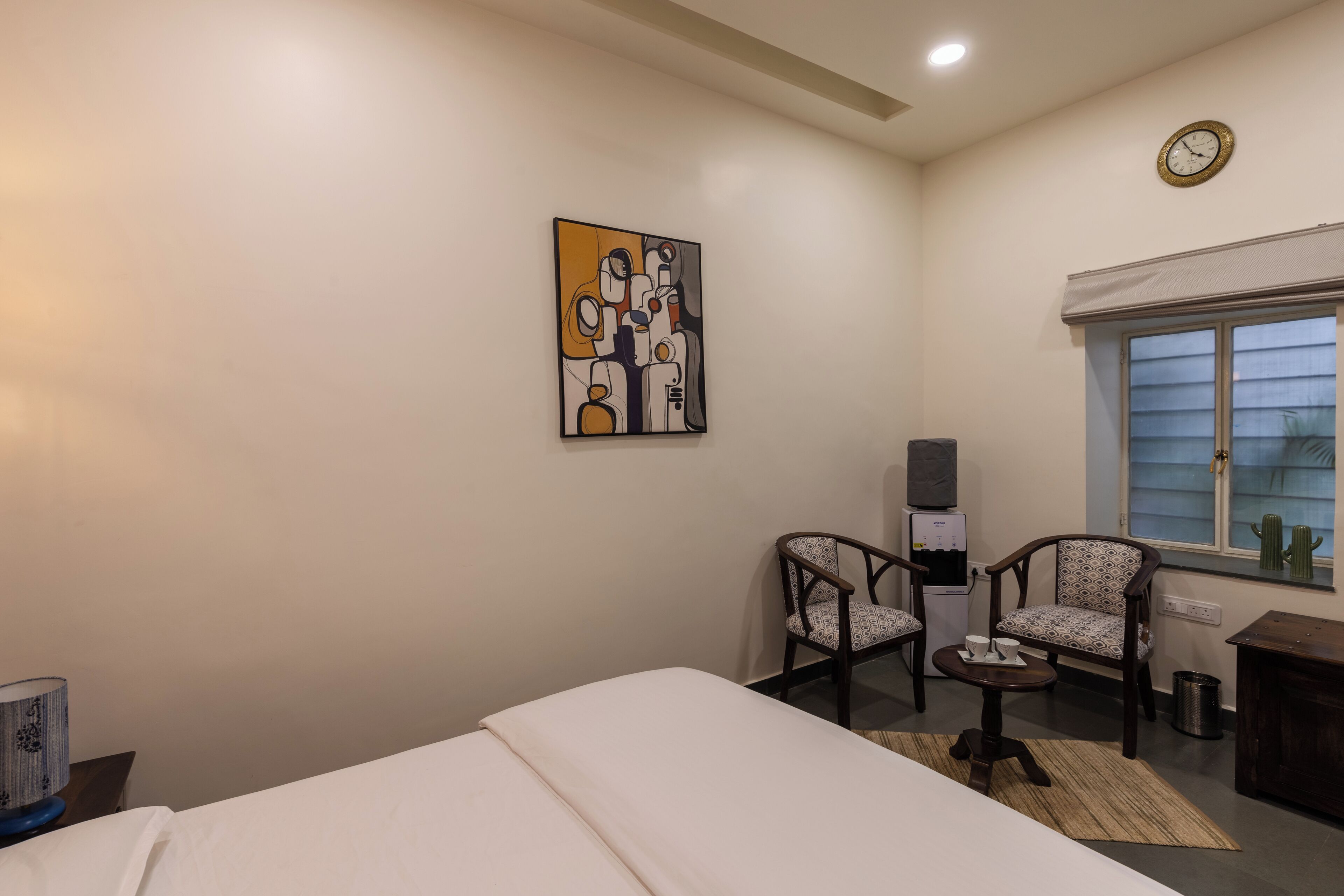 A hotel room with a bed, two chairs, a small table, a clock, and a window with a cactus decoration.
