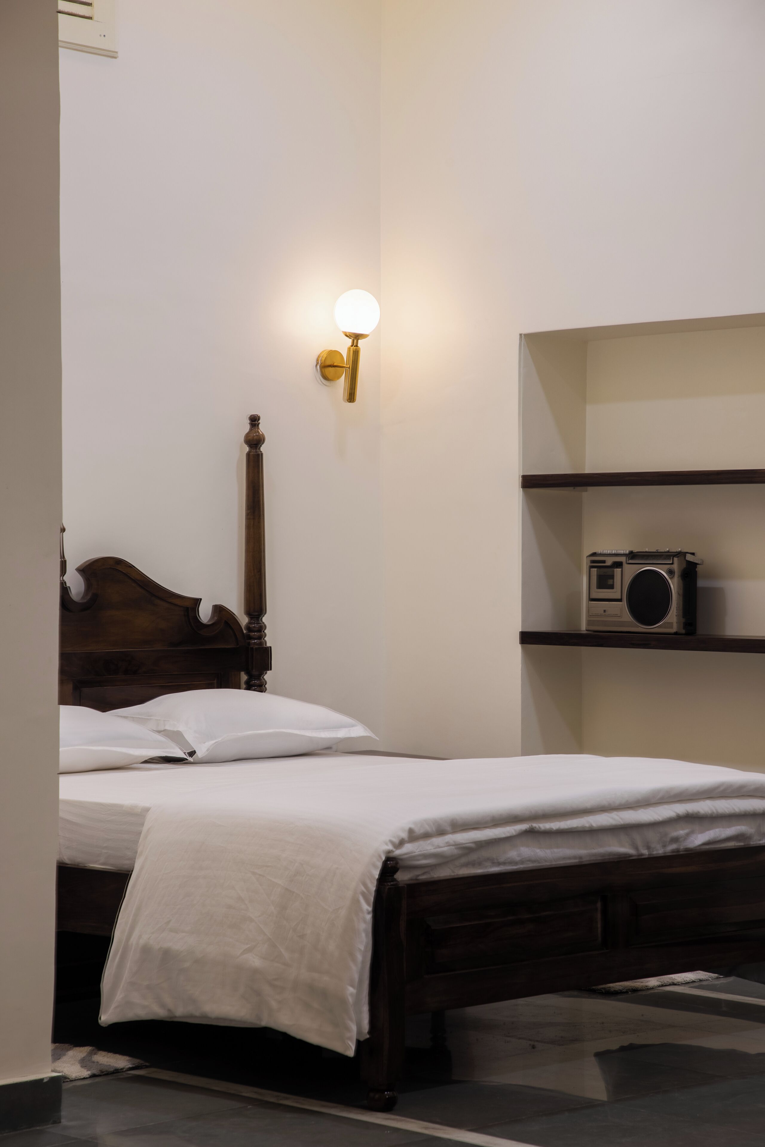 Un dormitorio con una cama de madera, ropa de cama blanca, una lámpara de pared y una estantería con un radio.
