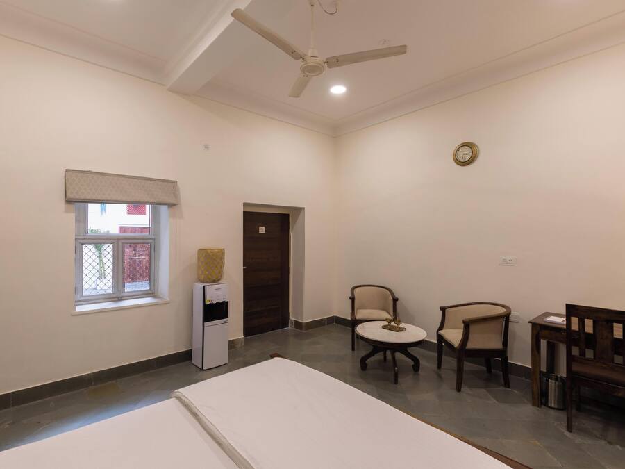 A hotel room with a bed, a ceiling fan, a small table with chairs, a refrigerator, and a window with a view.