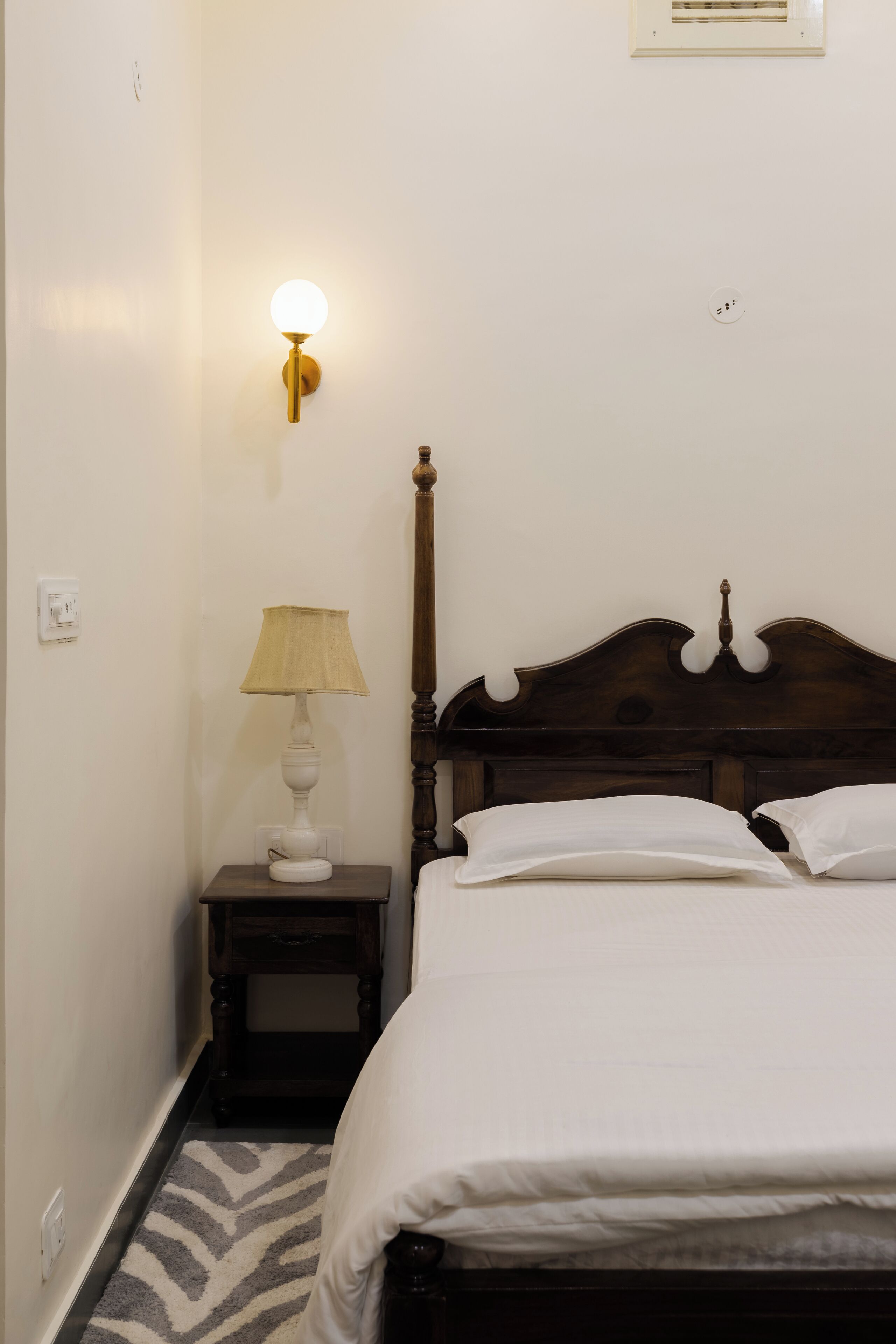 Una cama bien tendida con ropa blanca en una habitación con cabecera de madera. Hay una mesita de noche con una lámpara y un aplique de pared.