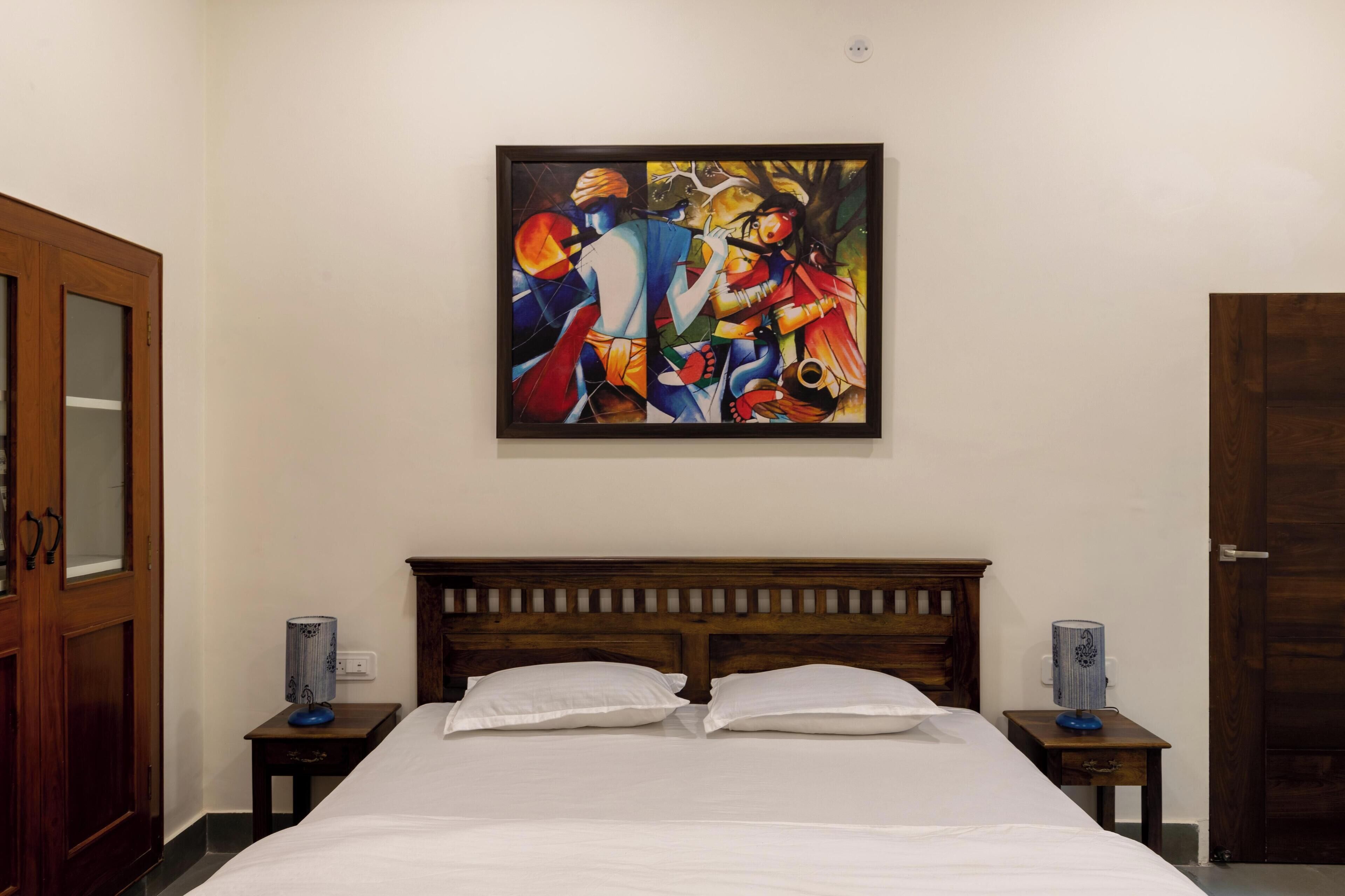 Een slaapkamer met een houten hoofdbord, een bed met witte lakens, twee nachtkastjes met lampen, en een kleurrijk abstract schilderij aan de muur.
