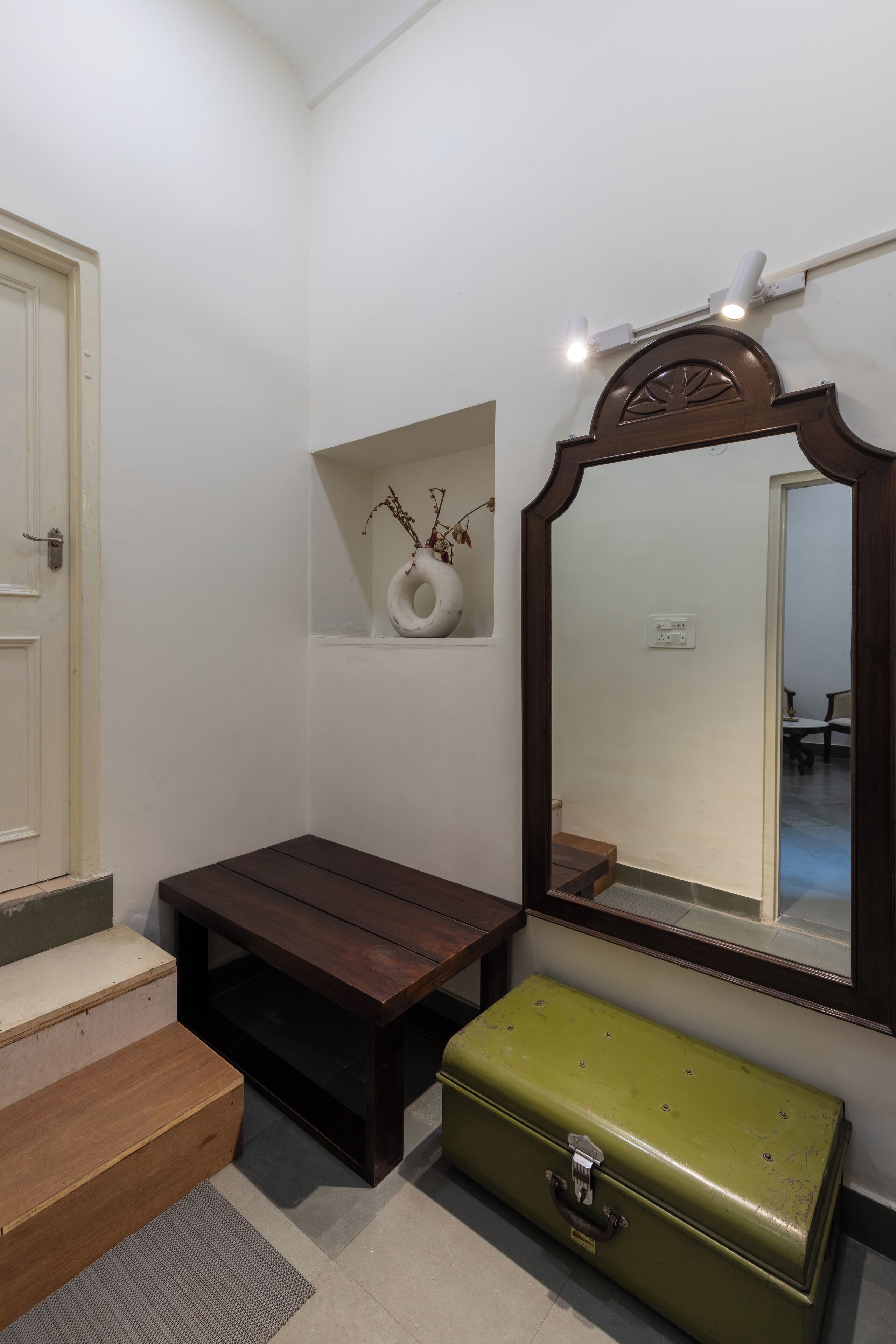 Um quarto pequeno com um banco de madeira, uma mala verde, um vaso branco com galhos secos e um espelho com moldura de madeira.