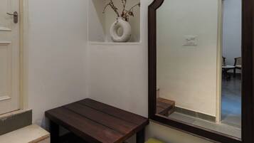Um quarto pequeno com um banco de madeira, uma mala verde, um vaso branco com galhos secos e um espelho com moldura de madeira.