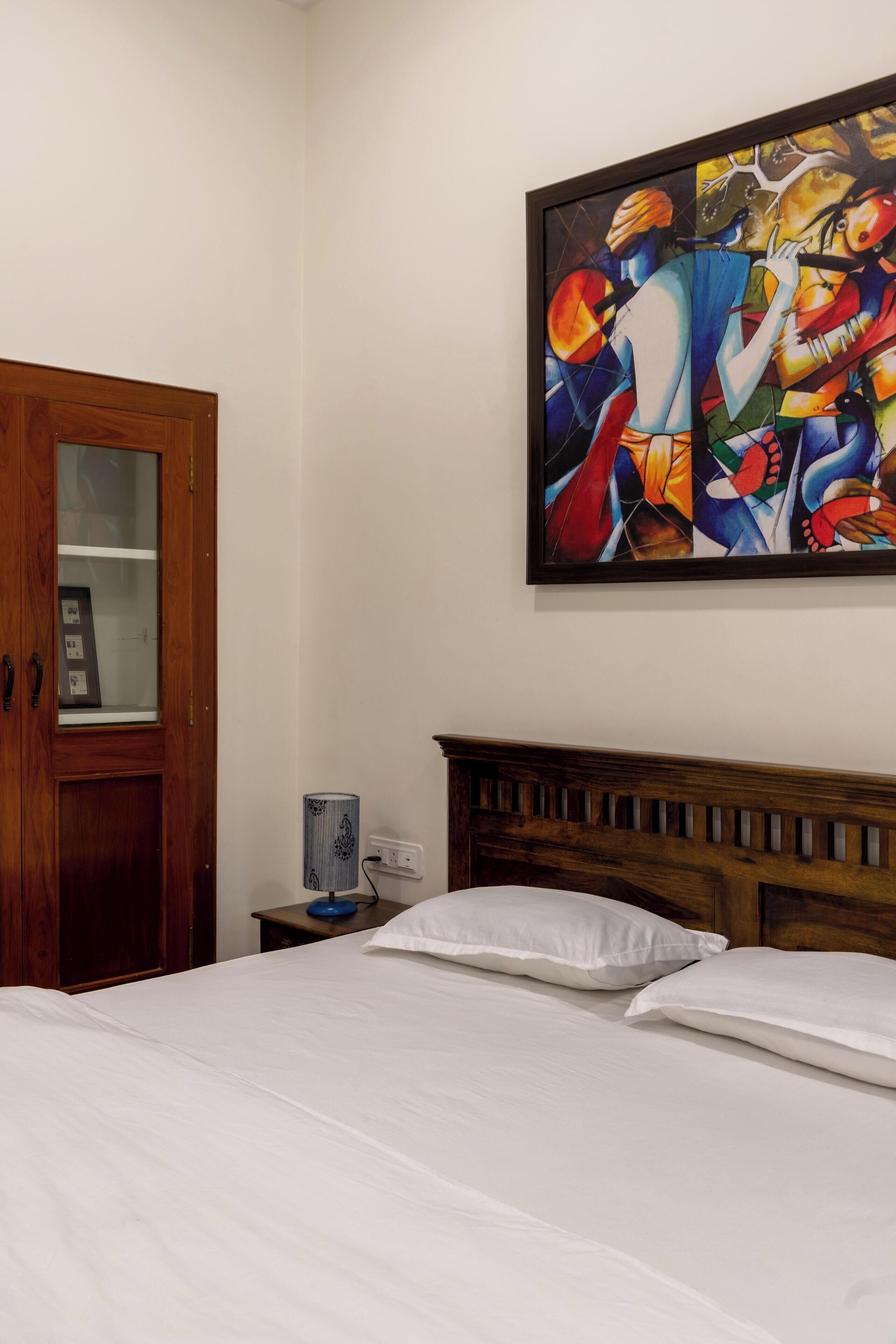 A bedroom with a wooden headboard, a bed with white linens, a nightstand with a lamp, and a wooden wardrobe with a glass door.