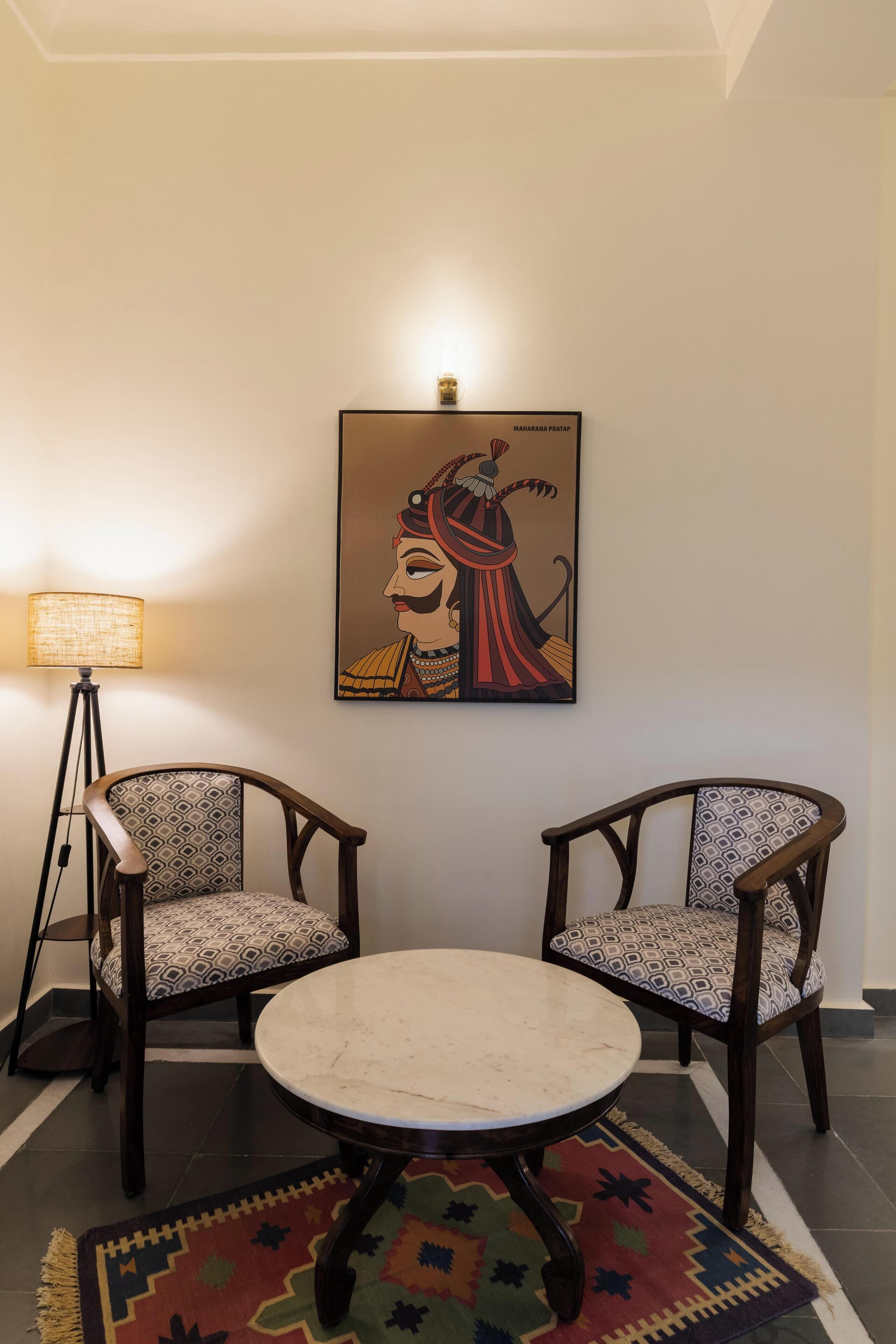 A room with two wooden chairs with patterned cushions, a round table, a colorful rug, and a wall-mounted lamp.