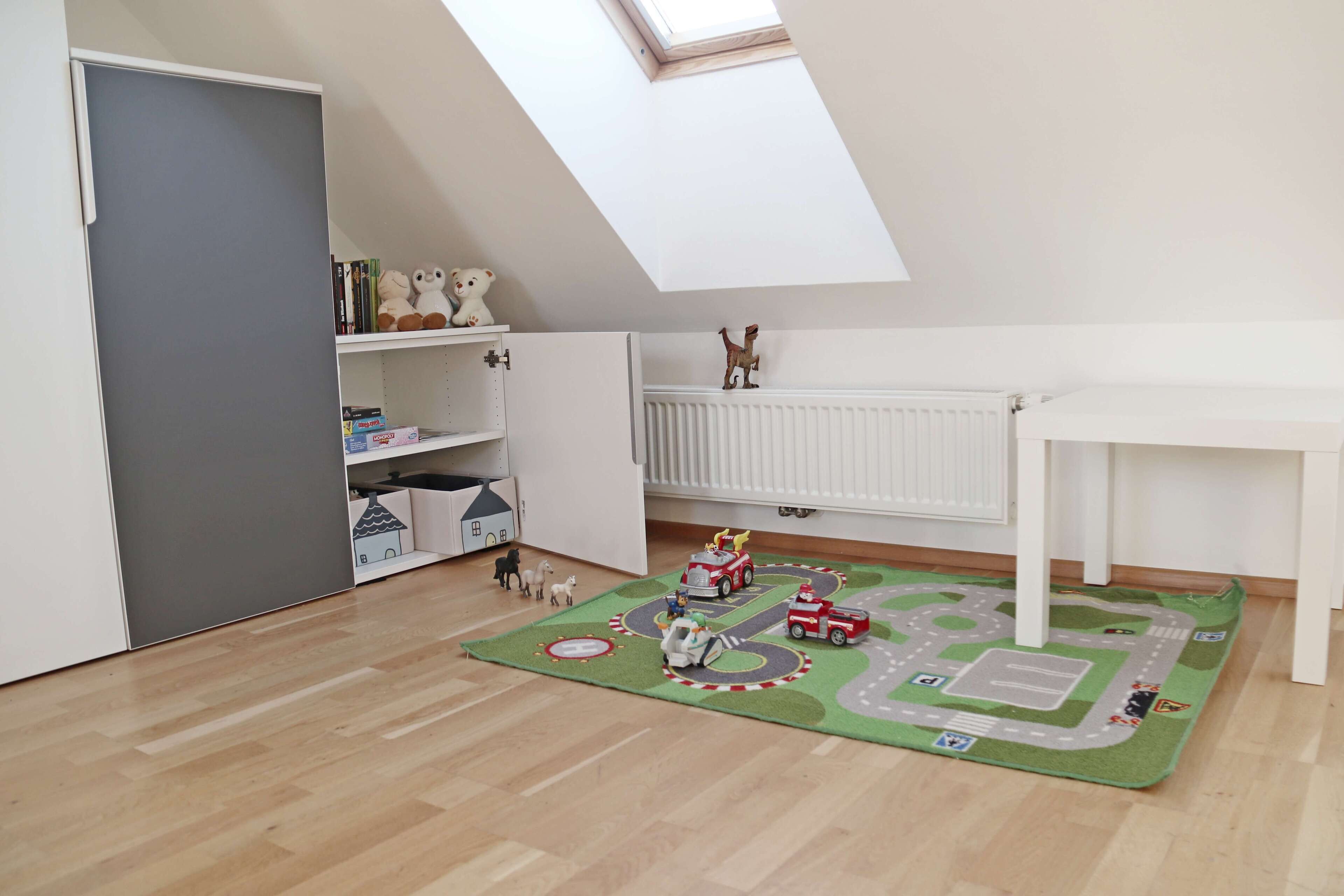 Una stanza da gioco per bambini con un velux, un tappeto giochi e macchinine, e un termosifone bianco.