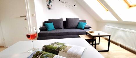 Un salon moderne avec un canapé gris, des coussins turquoise, une table basse et un magazine posé sur une table blanche. Un verre de vin rouge se trouve sur la table.