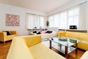 A modern living room with a glass coffee table, yellow sofas, a white radiator, and a large window with sheer curtains.