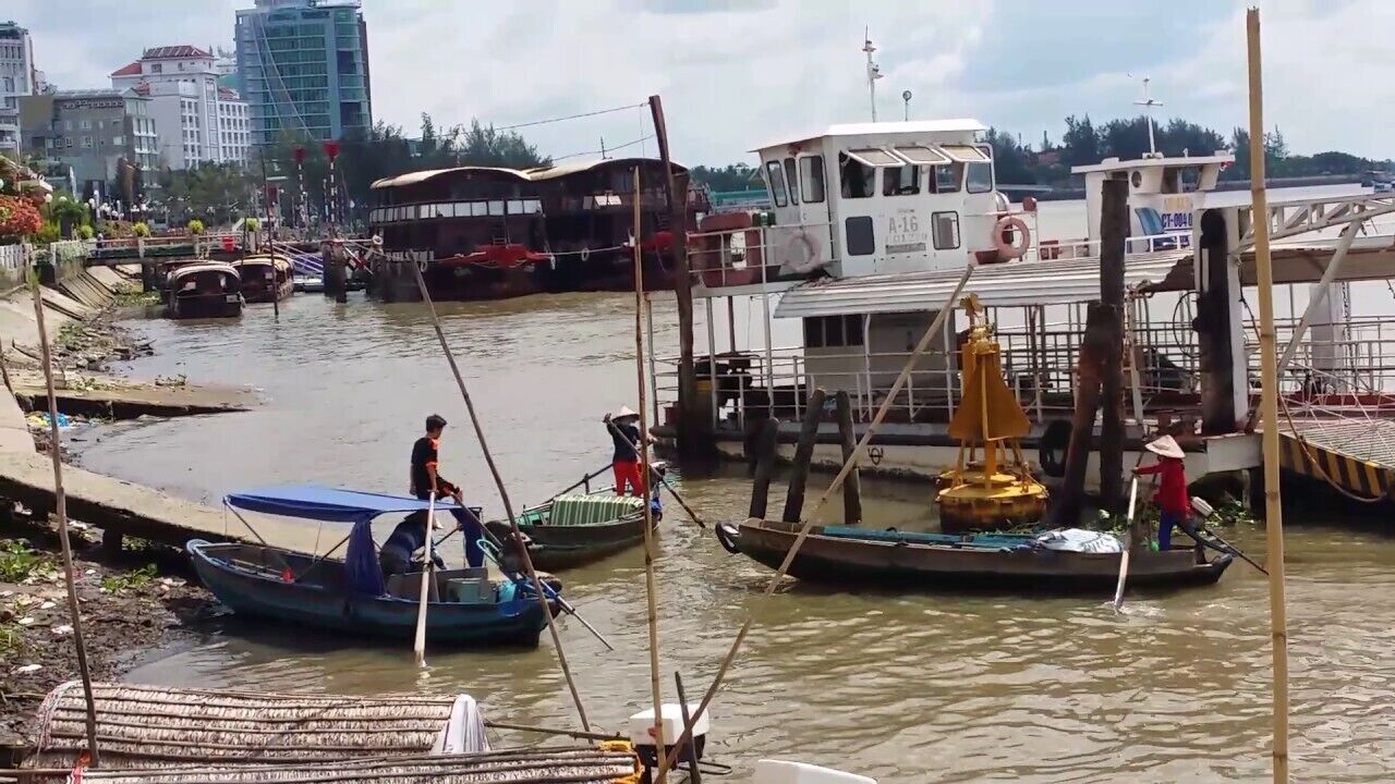 A riverfront with boats docked, a white boat with the number A-16, and a person in a red shirt standing on a dock.
