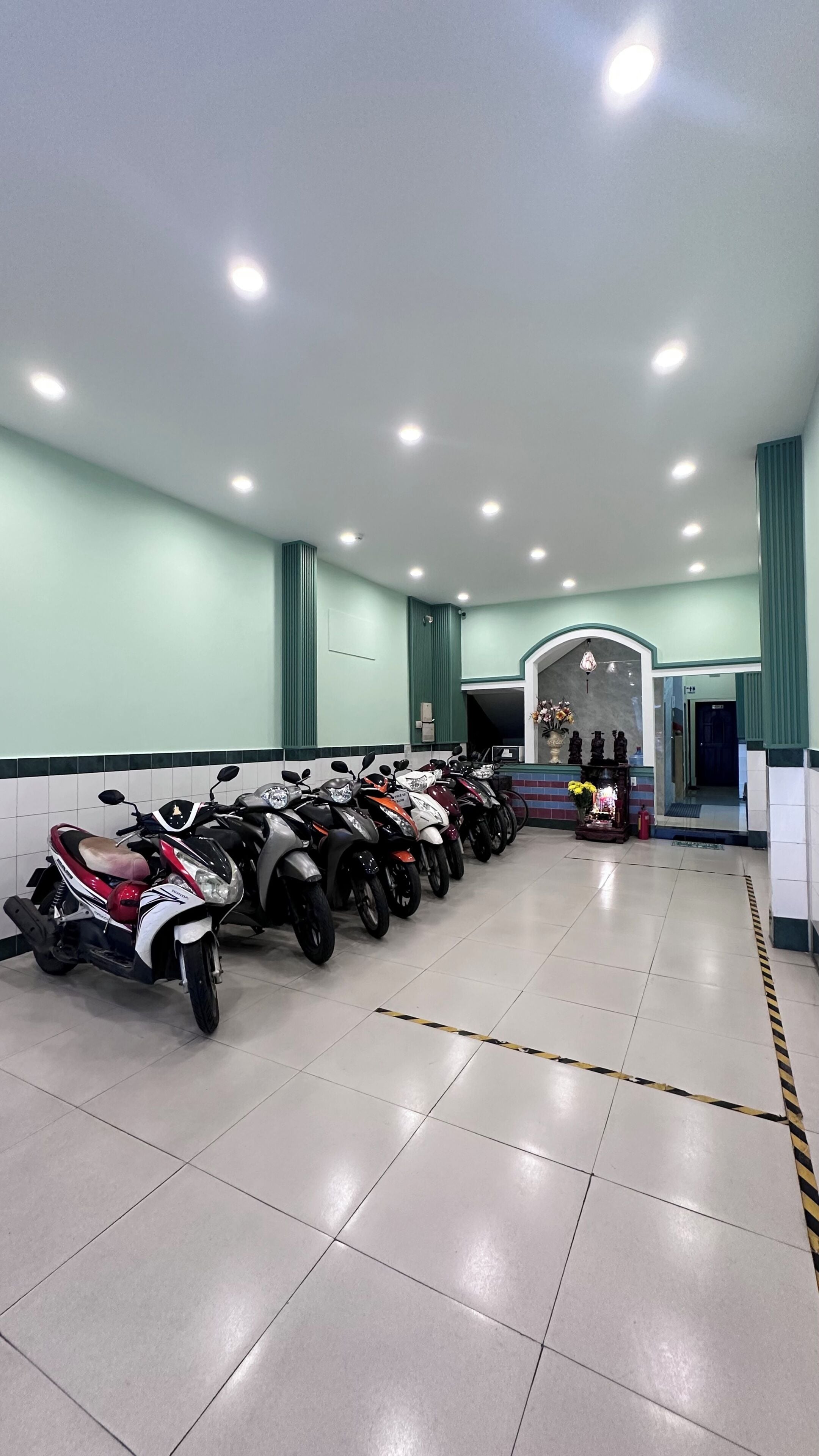 A clean, well-lit indoor space with a row of parked scooters, a tiled floor, and a reception area with a counter and decorative items.