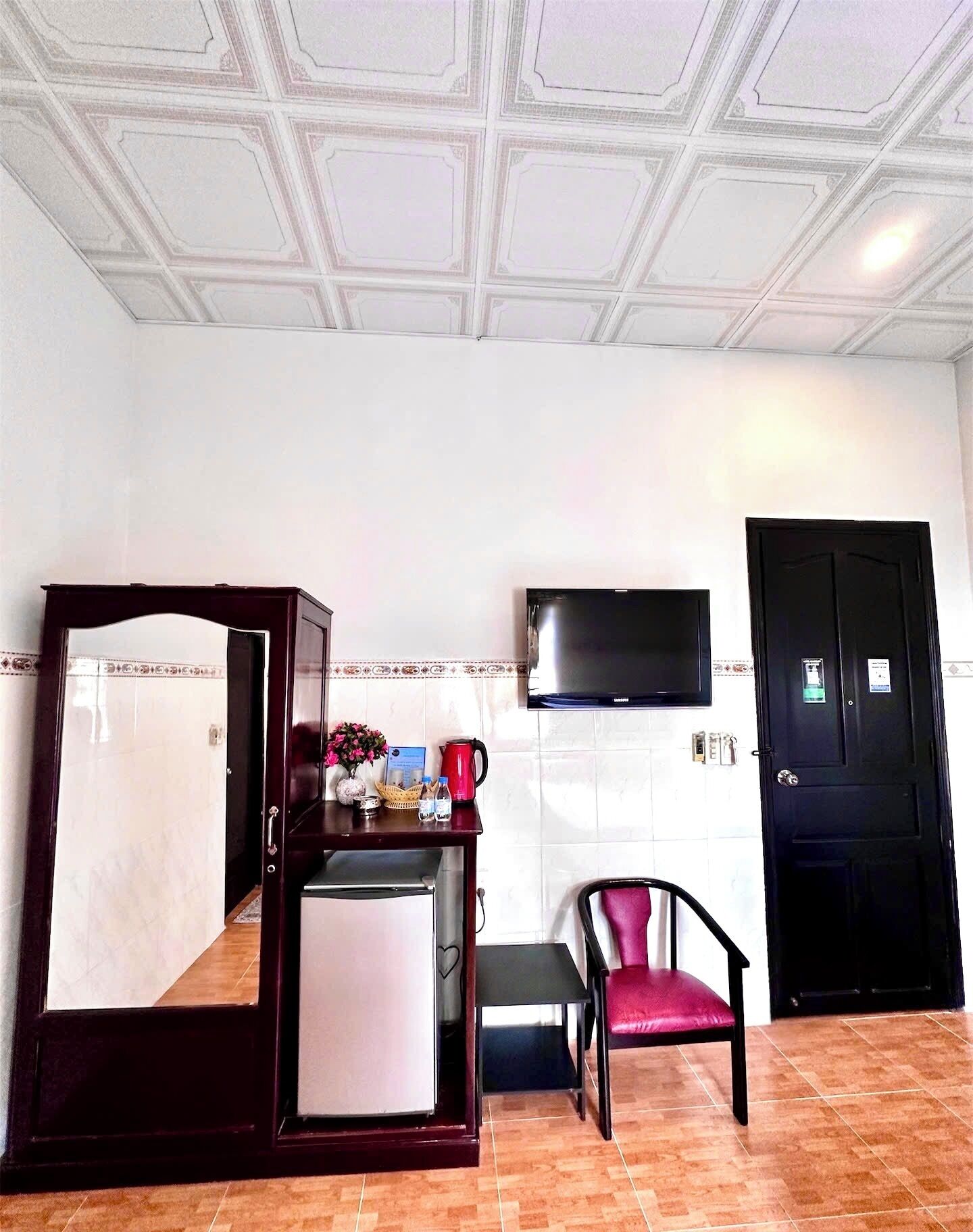 A room with a wooden four-poster bed, a small table with a vase and flowers, a black chair, a flat-screen TV mounted on the wall, and a black door.
