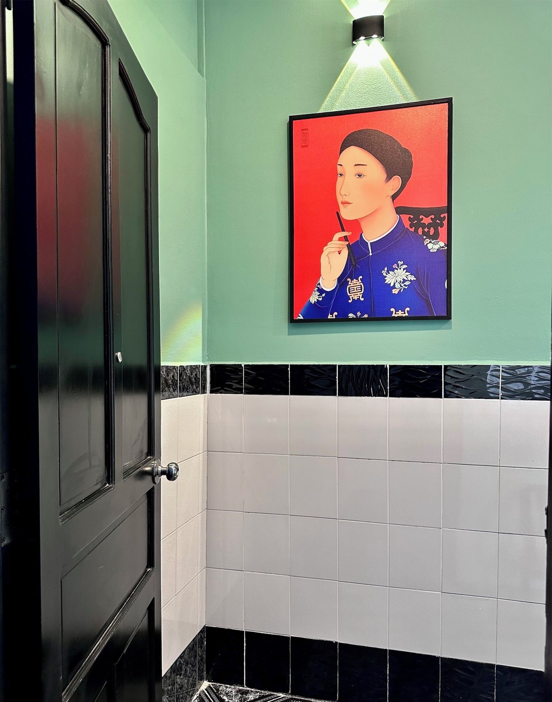 A bathroom with a black door, a wall-mounted light fixture, and a framed artwork above a tiled wall.