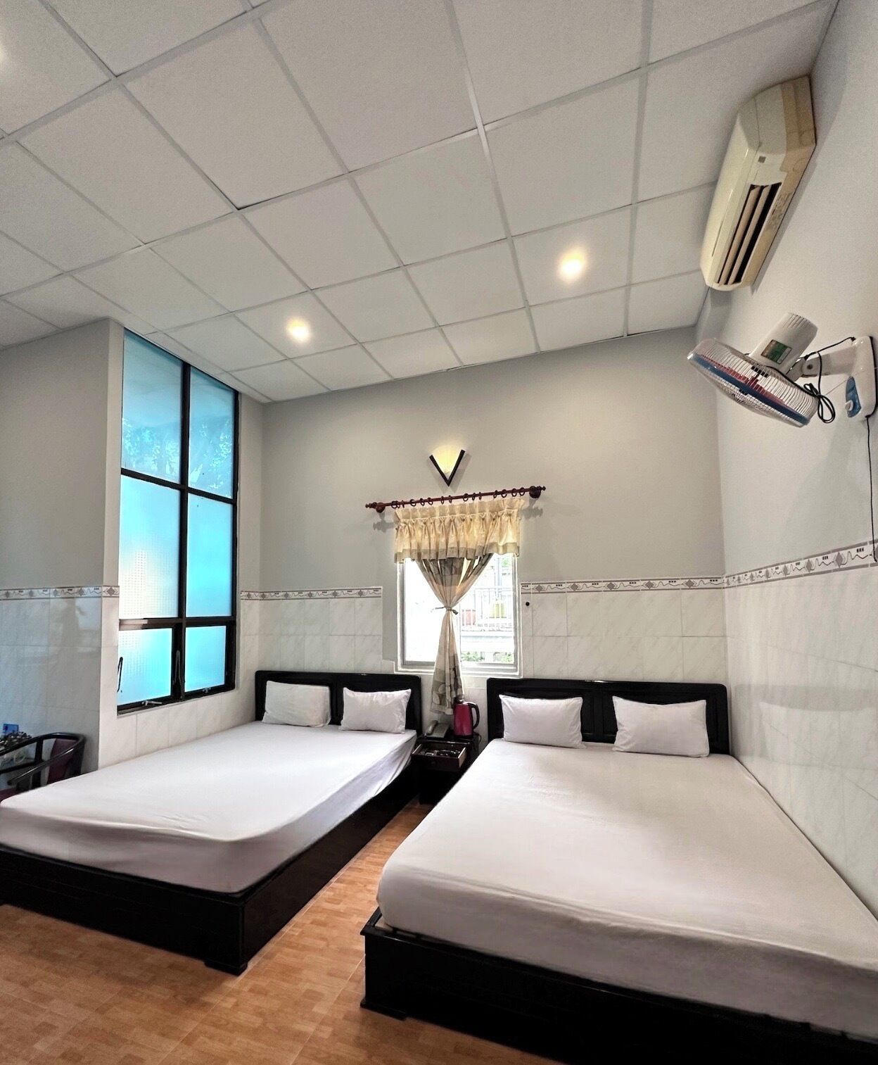 A hotel room with two beds, a window with a view, an air conditioner, and a bedside table with a lamp.